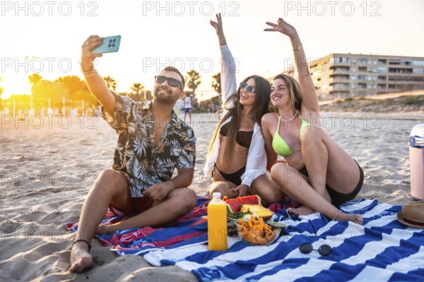 Three friends are sitting on a beach towel, taking a selfie while enjoying snacks and drinks during a vibrant sunset