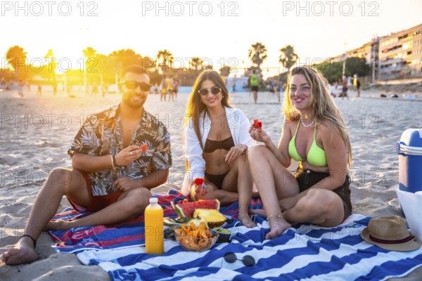 Three friends enjoying a picnic on the beach at sunset, eating watermelon and other fresh fruit
