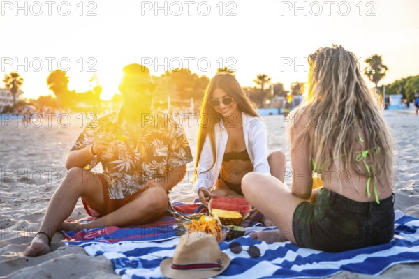 Group of friends enjoying watermelon and pineapple during a sunset picnic on a beach towel, creating a vibrant summer scene