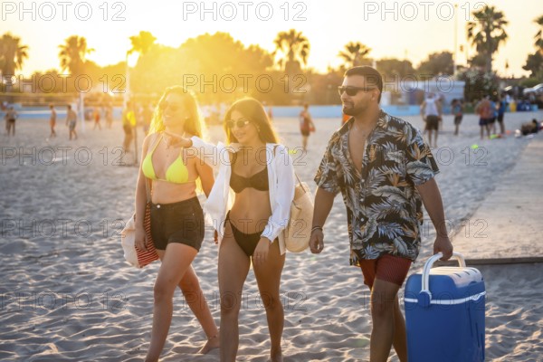 Three friends walking on the beach at sunset, one of them pointing towards something while another carries a cooler