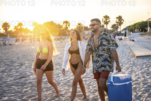 Three friends enjoying a leisurely stroll along the beach at sunset, carrying a cooler, ready for a fun evening