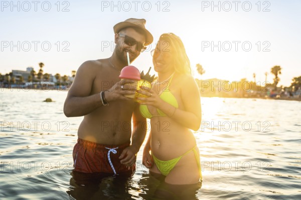 Happy couple enjoying refreshing cocktails while standing in the ocean during a beautiful sunset