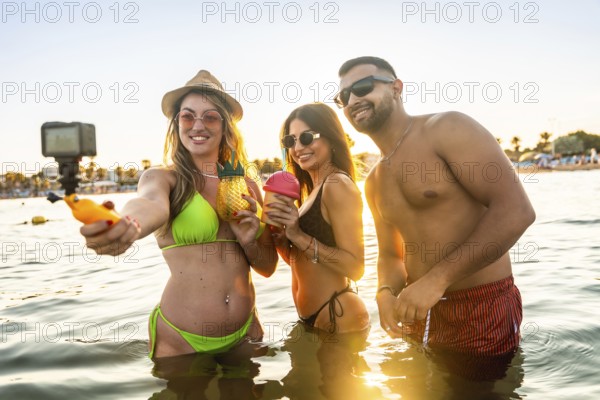 Young tourists capturing a joyful selfie in the sea during a vibrant sunset, celebrating summer vacation with friends and drinks