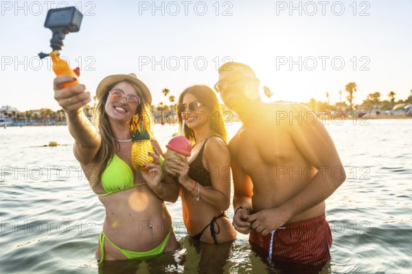 Group of cheerful young friends taking a selfie with action camera while enjoying cocktails in the sea at sunset