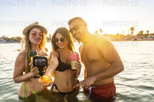 Three friends enjoying summer vacation, taking a selfie with action camera in the sea at sunset, holding tropical drinks