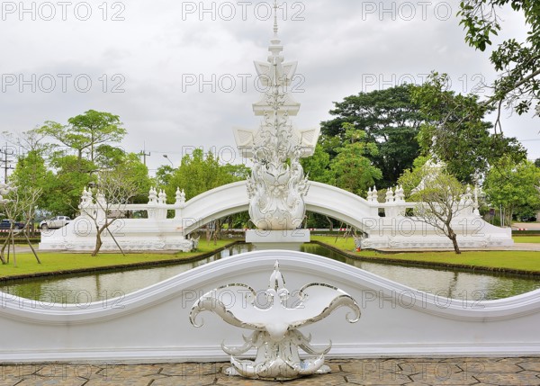 Ornate white bridge reflecting beautifully in the tranquil canal of a thai temple, surrounded by lush green trees under a cloudy sky