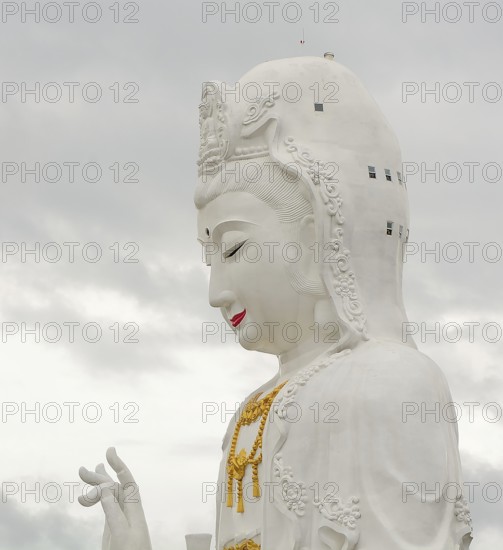 Majestic white guan yin statue, a prominent buddhist figure, stands tall against a cloudy sky in thailand, showcasing intricate details and serene expression