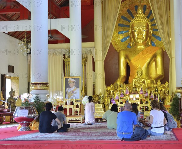 Tourists kneeling and praying in front of a giant golden buddha statue inside a buddhist temple in thailand, demonstrating reverence and cultural immersion