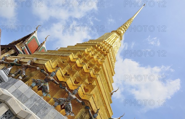 Golden chedi reaching for a bright blue sky, supported by decorative figures, within the majestic grand palace in bangkok, thailand