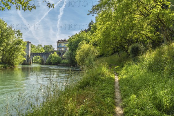 Lush vegetation frames a tranquil pathway meandering along the gave d'oloron river, leading towards the historic bridge and charming houses of sauveterre de bearn, france, under a vibrant sky