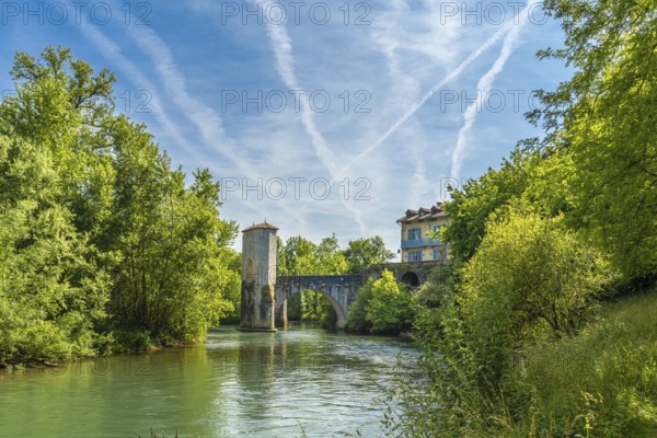 Scenic view of the historic stone bridge spanning the gave d'oloron river, with lush greenery and a picturesque building in sauveterre de bearn, france, under a vibrant sky with contrails