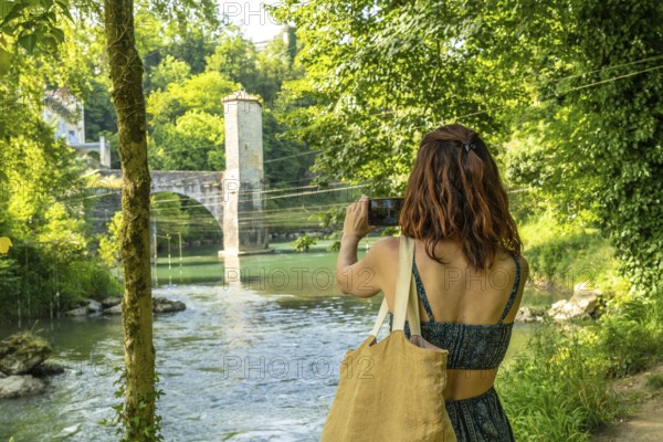 Young woman photographing a medieval bridge over gave d'oloron river in sauveterre de bearn, a picturesque village in the pyrenees atlantiques region of france