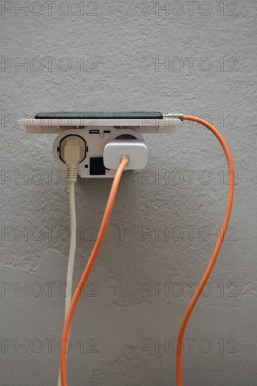 A smartphone is plugged into a wall outlet, showcasing cables in orange and white. This setup emphasizes the convenience of charging multiple devices in an indoor space, emphasizing technology use