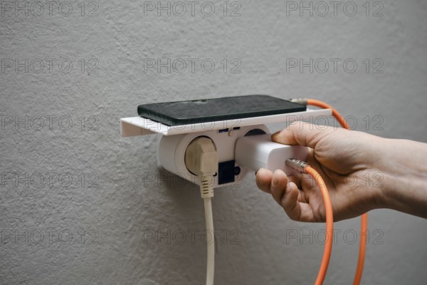 A person is holding a charger connector and plugging it into a wall-mounted power outlet. A smartphone is resting on a shelf above, connected by a charging cable