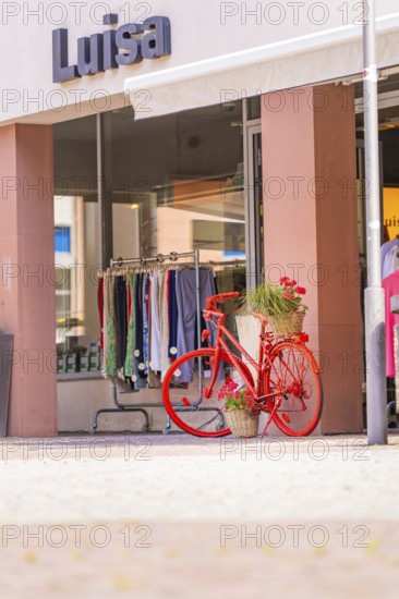 Red bicycle with flower baskets in front of a clothing shop in a busy shopping street, small town of Perle Calw, Black Forest, Germany
