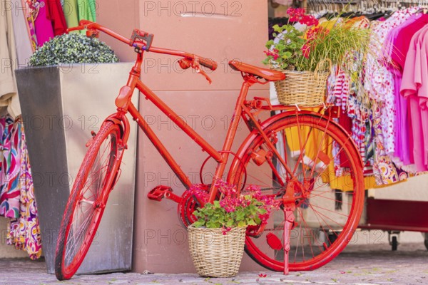 A decorated red bicycle stands in front of a clothing shop with colourful clothes, small town of Perle Calw, Black Forest, Germany