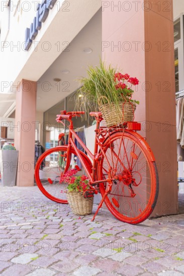A red bicycle with flower baskets stands decoratively on a pavement in front of shops, small town of Perle Calw, Black Forest, Germany