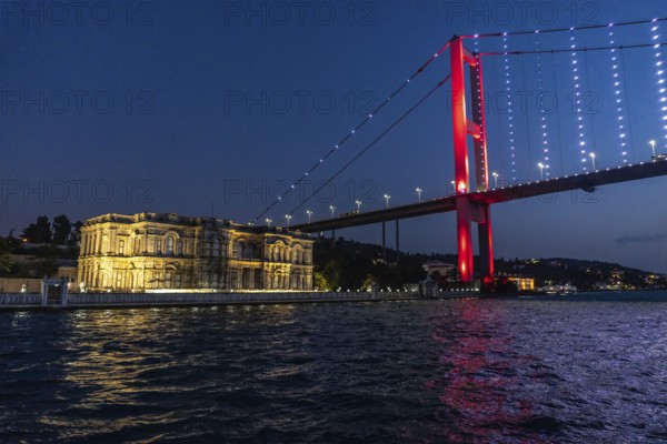 The illuminated suspension bridge over bosphorus strait paints the istanbul night sky red, creating a vibrant contrast with the historic palace