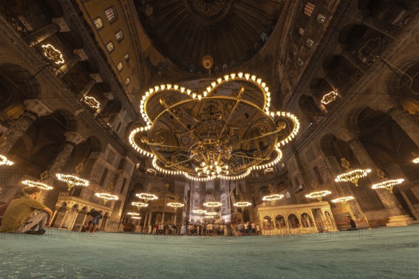 Tourists gather beneath the magnificent chandeliers within the hagia sophia, a historical landmark in istanbul, turkey, experiencing its architectural grandeur