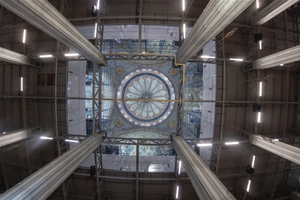 Wide angle view capturing ongoing restoration efforts within a mosque, highlighting scaffolding encasing a beautifully decorated dome, showcasing intricate details and the scale of the project