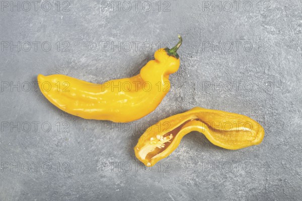 Two unusual yellow peppers are displayed on a textured gray surface. One pepper is whole while the other is cut in half, revealing its inner seeds and bright flesh, highlighting their unique shapes