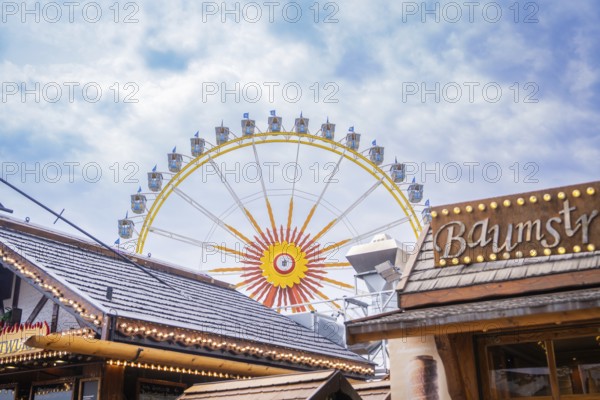 Large Ferris wheel behind wooden buildings, in a bright and cloudy sky, Olympiapark, Munich, Germany
