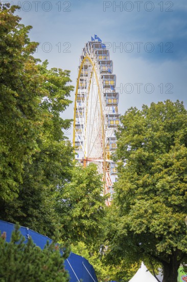 Ferris wheel hidden behind dense green trees under a blue sky, Olympiapark, Munich, Germany