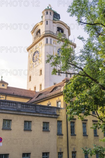 Historic clock tower towers over a yellow building, surrounded by green nature in bright sunshine, Munich, Germany
