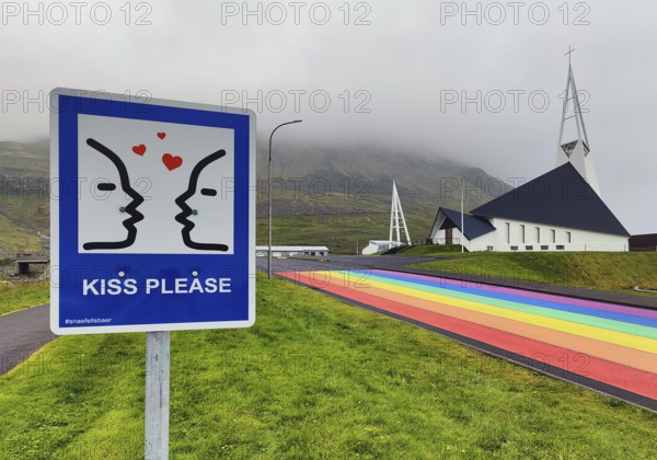 Sign with the invitation to kiss with a street painted in the colours of the rainbow and the church, Ólafsvík, Snæfellsnes, Iceland