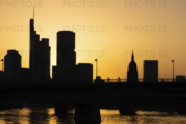 Two cyclists pass the rafter bridge over the Main in the evening while the sun sets behind the Frankfurt banking skyline, Frankfurt am Main, Hesse, Germany