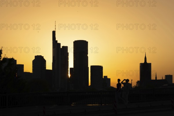 Two men cross the rafter bridge over the Main in the evening as the sun sets behind the Frankfurt banking skyline, Frankfurt am Main, Hesse, Germany