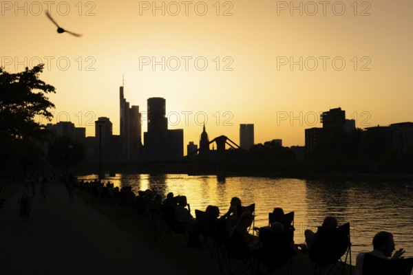 People sitting on the banks of the Main in the evening while the sun sets behind the Frankfurt banking skyline, Frankfurt am Main, Hesse, Germany