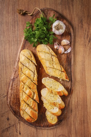 Garlic bread, baguette with garlic butter and herbs, on a wooden board, with herbs and garlic cloves
