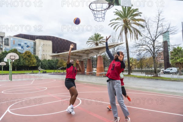 Two diverse women in red jerseys actively playing basketball on an outdoor court. Reaching for the ball near the hoop. Promoting sport. Healthy lifestyle. And multicultural engagement in an urban park