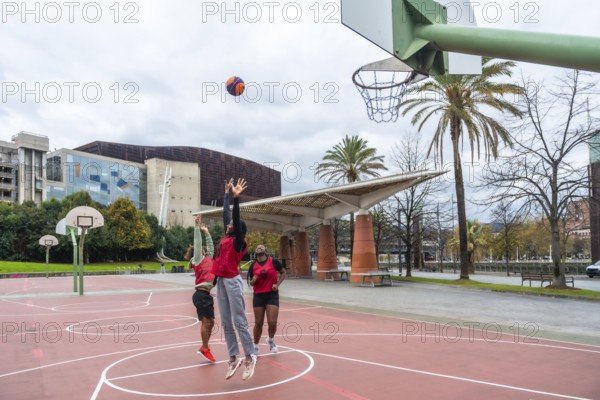 Diverse group of friends enjoying a casual game of street basketball on a red court, actively jumping and attempting to score under a cloudy sky in a modern urban environment