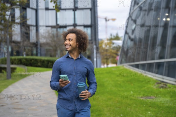 Smiling young Mixed-race man with curly afro hair walking on a path in a modern city park, holding a smartphone and a portable reusable cup, looking to the side