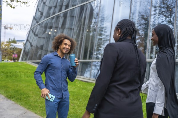 Diverse colleagues are enjoying a casual coffee break outdoors, talking and smiling against a backdrop of modern glass architecture, fostering connection and teamwork