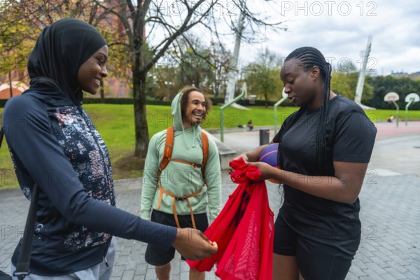 Three diverse friends, including two women wearing sportswear, are cheerfully gathering their gear on a basketball court, representing community, modern lifestyle, and friendship