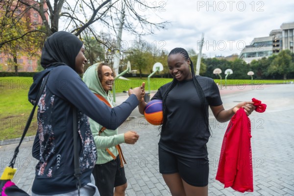 Three diverse friendswoman in hijab, man with curly hair, and woman holding a colorful basketballcheerfully high five on an outdoor urban court, celebrating sport and friendship