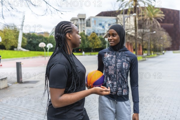 Diverse young women, one wearing a hijab and another with braids, talking and bonding while holding a basketball outdoors in an urban basketball court setting