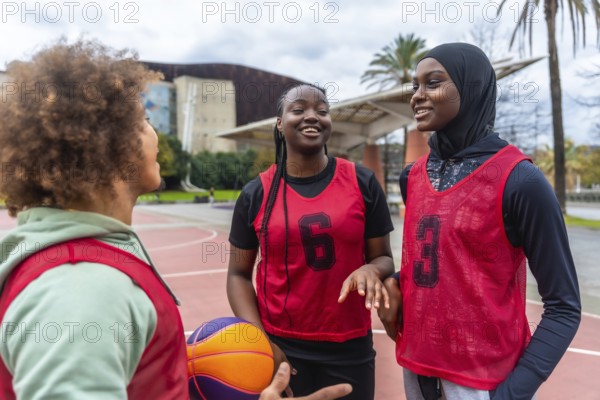 Three diverse young women, including one wearing a hijab, smiling and discussing before playing basketball on an outdoor urban court, representing teamwork and friendship