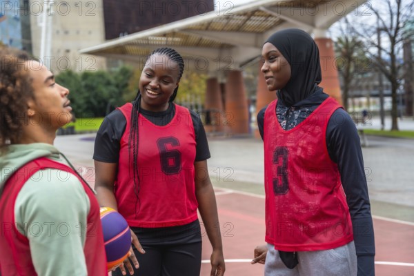 Group of diverse young athletes, including men and women, wearing red training bibs and a hijab, smiling and talking on an urban basketball court after playing a game