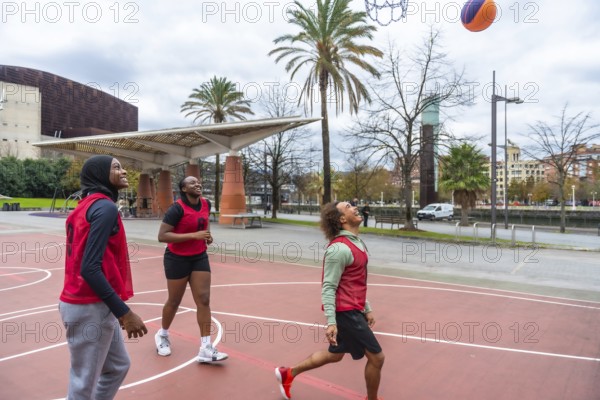 Diverse group of young adult friends playing basketball on an urban court, with a woman wearing a hijab actively participating and all looking up as the ball approaches the hoop