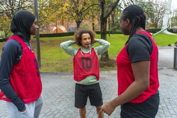 Group of diverse young adults wearing red training bibs, including a woman in a hijab, standing in a park, actively communicating and discussing while preparing for a sporting activity