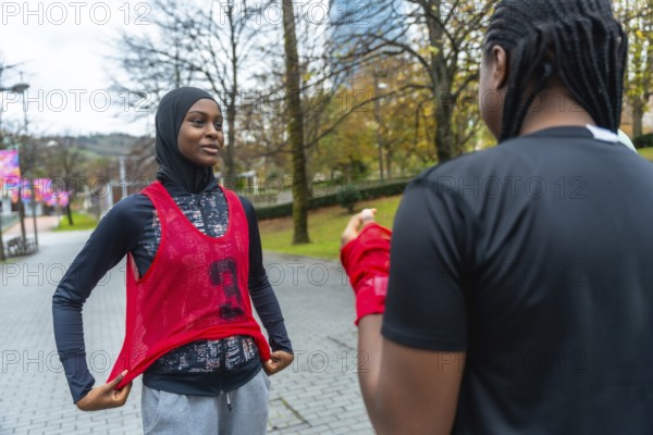 Young black woman wearing a hijab and training bib talking with a friend while preparing for her workout in an urban park, embodying fitness and diversity concepts