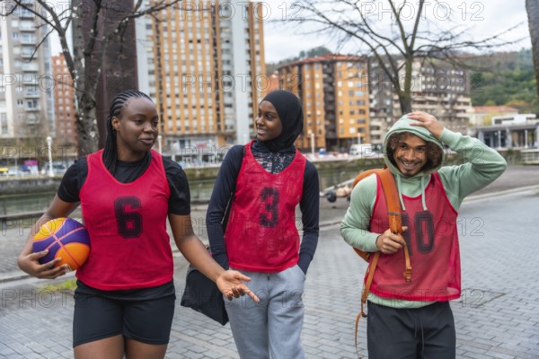 Diverse friends in sportswear and bibs walking and conversing on an urban street, carrying a basketball after playing a game, representing teamwork and friendship