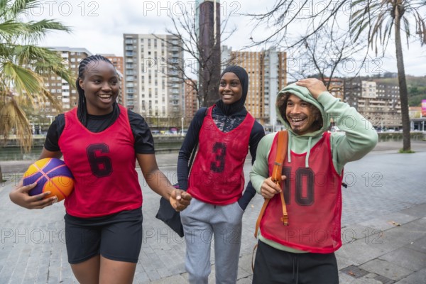 Three diverse friends, a young woman holding a basketball and two people wearing red bibs, walking and smiling against an urban background, symbolizing friendship and sport