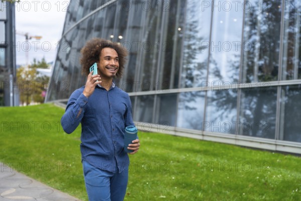Smiling young Mixed-race man with curly hair walking confidently along a modern office building, holding a reusable cup and having a business conversation on his smartphone