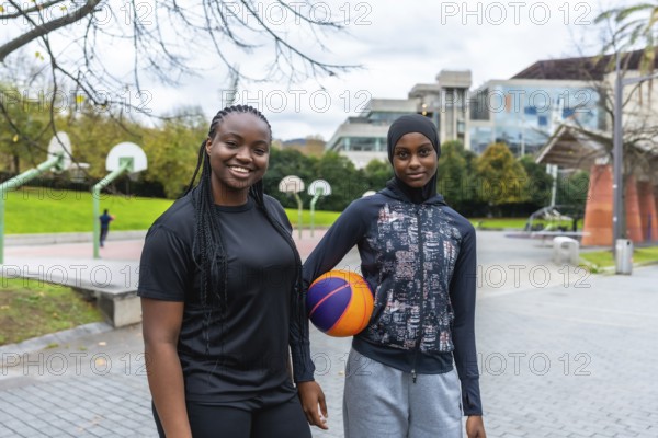 Two diverse young women, one wearing a hijab, smiling and holding a basketball while standing on an outdoor park court, representing athleticism, friendship, and urban sport