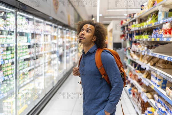 Young man with orange backpack studying refrigerated dairy products on supermarket shelves while walking the modern grocery aisle, contemplating choices during routine shopping trip
